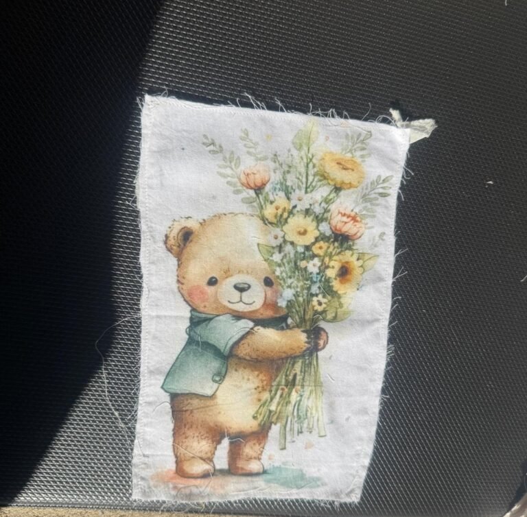 This appears to be a printed fabric patch featuring a cute illustration of a teddy bear holding a bouquet of flowers.